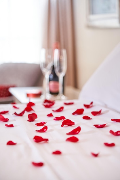 Closeup of comfortable bed with scattered red roses petals and bottle of champagne with glasses placed on bedside table in hotel room prepared for honeymoon of couple