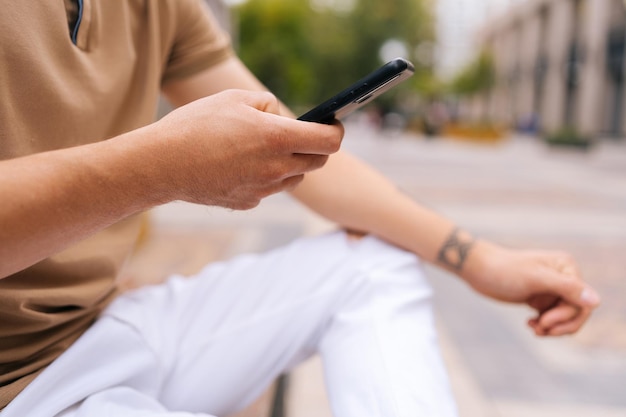 Closeup cropped shot of unrecognizable tattooed man sitting on bench using mobile phone looking to screen on city street