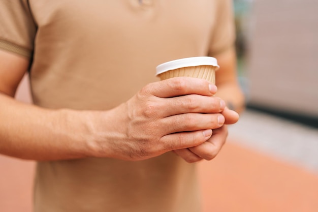 Closeup cropped shot of unrecognizable young man wearing tshirt holding in hands cup with takeaway coffee to go standing posing with on city street