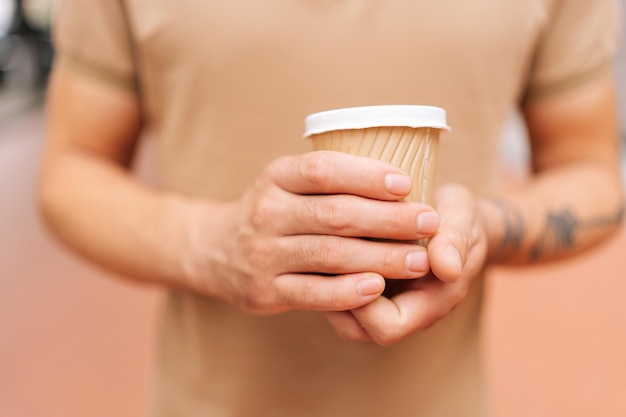 Closeup cropped shot of unrecognizable young man with tattooed arm wearing tshirt holding in hands cup with takeaway coffee to go