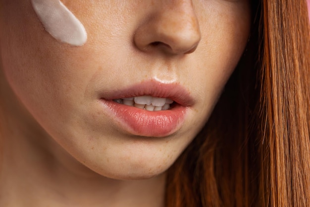 Closeup cropped shot of young caucasian redhead woman with freckles applying face primer or moisturizing, nourishing cream on her face. Beauty product, cosmetic, skin care.