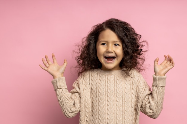 Closeup of cute surprised little brunette girl with curly hairstyle, raising hands with excited, amazed expression, screaming with delight