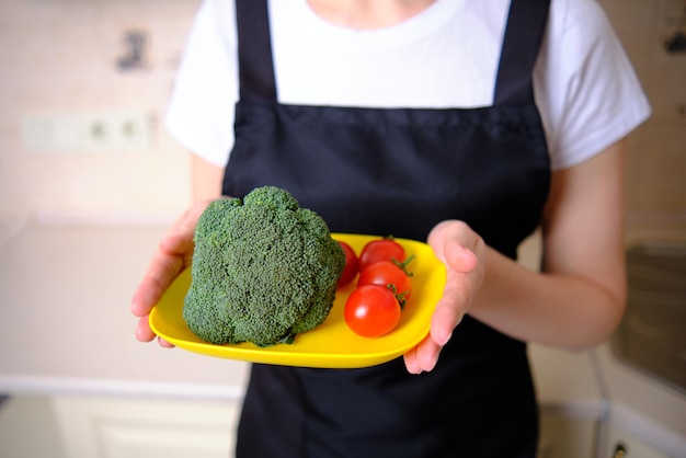 Closeup of a dish with broccoli and tomatoes in the hands of a young girl in a black apron