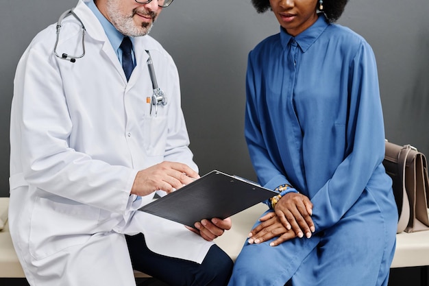 Closeup of doctor in white coat pointing at document and asking questions to patient at consultation