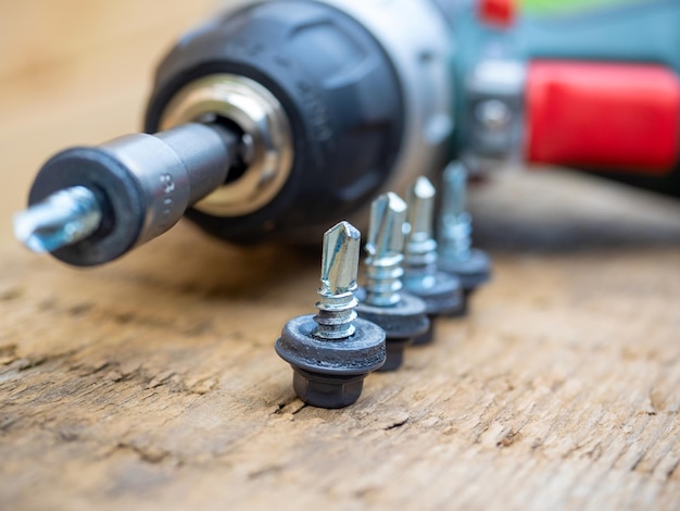 A closeup of an electric screwdriver with a nozzle on lies on a wooden background Next to it roofing screws on metal are laid out in a row The concept of repair and construction