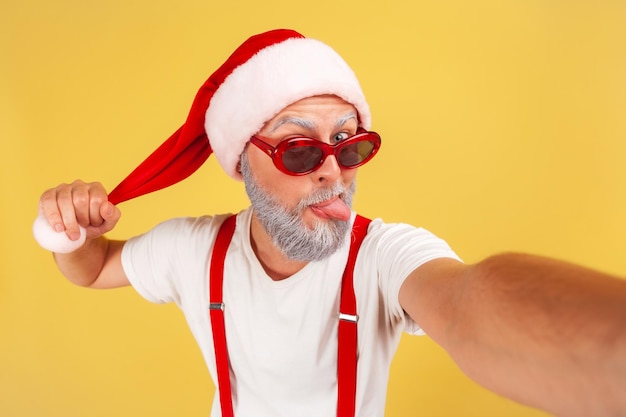 Photo closeup excited gray bearded man in red santa claus hat fooling around showing tongue out making selfie on camera keeping blog indoor studio shot isolated on yellow background