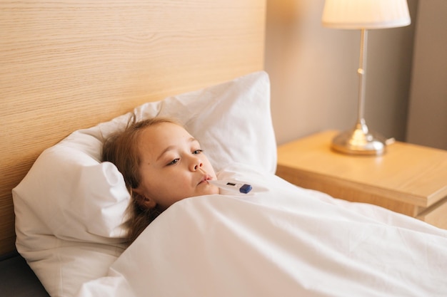 Closeup face of little sick girl lying in bed measuring temperature with thermometer in mouth at home