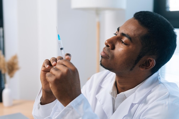 Closeup face of pensive black male doctor in white uniform thoughtful looking to syringe with vaccine in hand sitting at desk in medic clinic