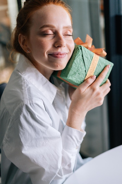 Closeup face of pretty redhead young woman gently cradling surprise gift box to face at home