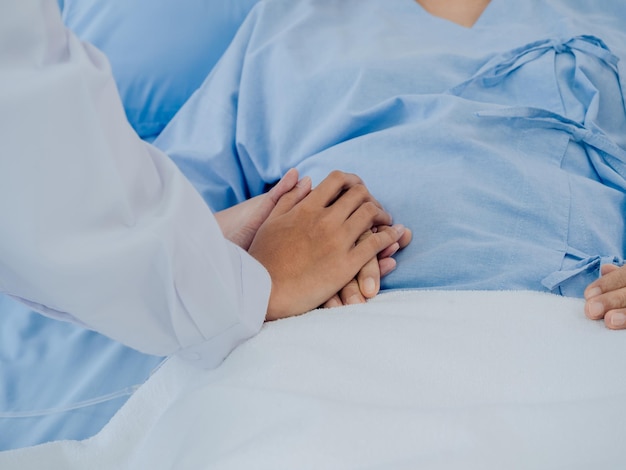 Closeup female doctor in white suit is holding on to elderly senior woman patient's hand in light blue dress with a saline hose lying on hospital bed encouraging the patient concept