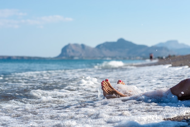 Closeup of female feet with pink pedicure in a foamy sea of a wave on pebble beach of the island Rhodes.