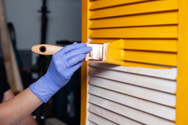 Closeup female hand in purple rubber glove with paintbrush painting natural wooden door with yellow paint.  creative design house interior. How to Paint Wooden Surface. Selected focus