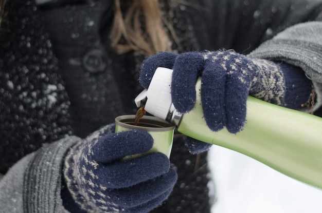 Closeup of female hands in knitted gloves pouring hot coffee from a thermos on a winter cold snowy day