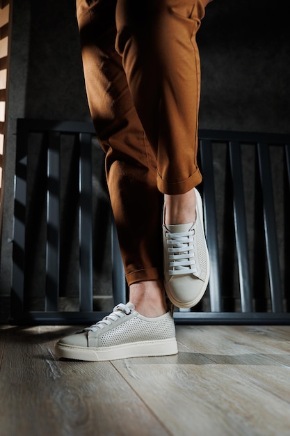 Closeup of female legs in leather white shoes Young woman standing in leather stylish white sneakers in brown pants Summer women's shoes Casual street style