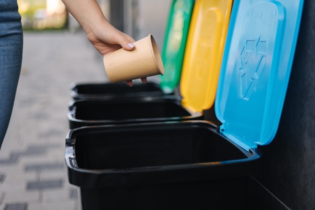 Closeup of female throwing cardboard cup in recycling bin different colour of recycling bins