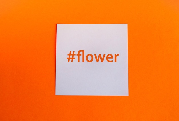 Closeup of a flower of the word flower with a hashtag on a white square piece of paper on an orange background flat lay