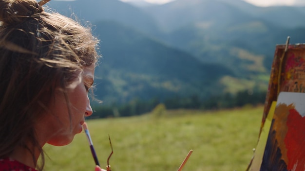 Closeup focused woman painting mountains scenery at summer holiday Portrait of talented girl looking to sketch standing green meadow Inspired artist drawing creative picture on easel outdoors