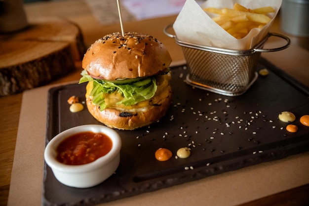 Closeup of fresh burger with basket of French fries on stone table with bowl of tomato sauceTasty delicious beef burger with fresh vegetables fries and coleslaw