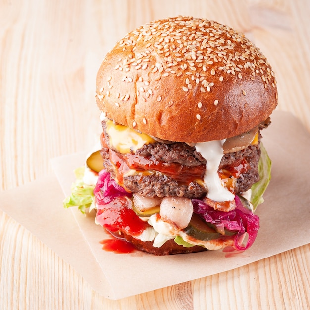 Closeup of a fresh sesame burger on white wooden board with ketchup and white sauce, cheese, mushrooms and red onion. Square shot