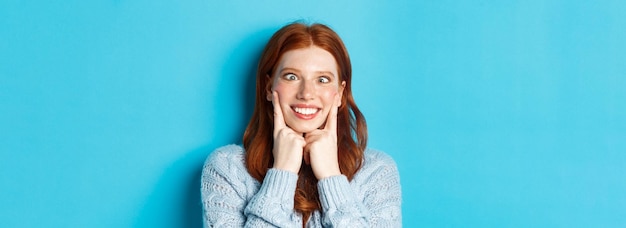 Closeup of funny redhead teen girl making faces squinting and squeezing cheeks standing against blue