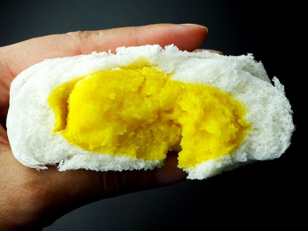 Closeup half inside of yellow custard cream bun in hand fingers on black background, yummy milk custard bun