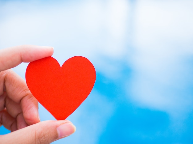 Closeup hand holding red paper heart shape on blue background