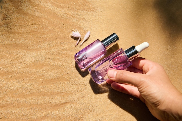 Closeup of the hand putting skin care beauty product packaging in a light violet glass bottle on golden sand background