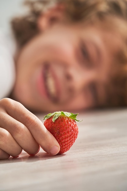 Closeup of hand of smiling child resting head on floor and looking at sweet berry at home on weekend day