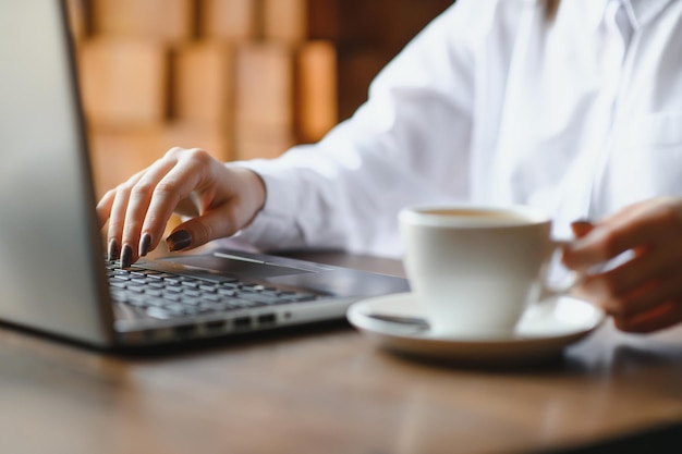 Closeup hands of business woman in pastel pink shirt working on laptop and making notices in her notebook on wooden table with a cup of coffee