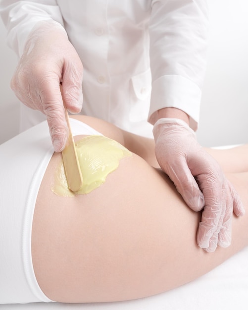 Closeup hands in gloves applies green hot wax on buttocks of woman using spatula Waxing procedure