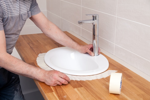 Closeup hands of a professional plumber worker installs a white oval ceramic sink on a wooden tabletop in the bathroom with beige tile, paste over sink with masking tape for applying sealant