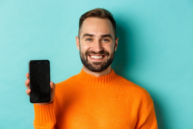 Closeup of handsome bearded guy in orange sweater showing smartphone screen and smiling showing prom...