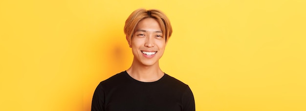 Closeup of handsome stylish asian guy with fair hair smiling happy over yellow background