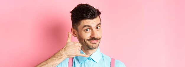 Closeup of handsome young guy with moustache showing phone gesture asking to call him standing on pi