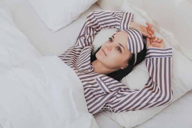 Closeup of happy and pleased smiling girl in striped pajamas and sleeping mask lying in bed on pillow enjoying the rest after a good healthy sleep resting in the morning in a good mood