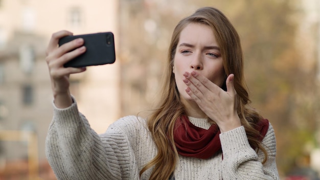 Closeup happy woman making selfie photo by mobile phone outdoors Portrait of funny girl pulling faces to smartphone on city street Cheerful hipster girl posing for cellphone camera outside