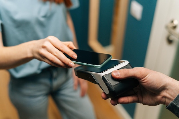 Closeup highangle view of unrecognizable courier male giving POS wireless terminal to making contactless payment using mobile phone to young female customer