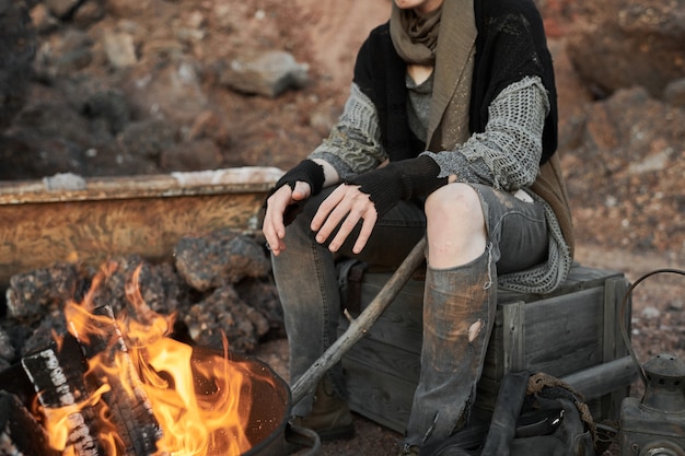 Closeup of homeless woman in torn clothes warming up near the fire outdoors