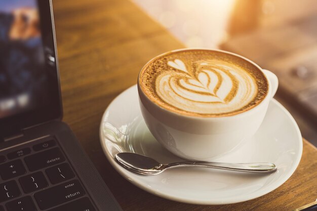 Closeup of Hot coffee latte with latte art milk foam in cup mug and laptop computer and smartphone on wood desk office desk in coffee shop at the cafeduring business work concept