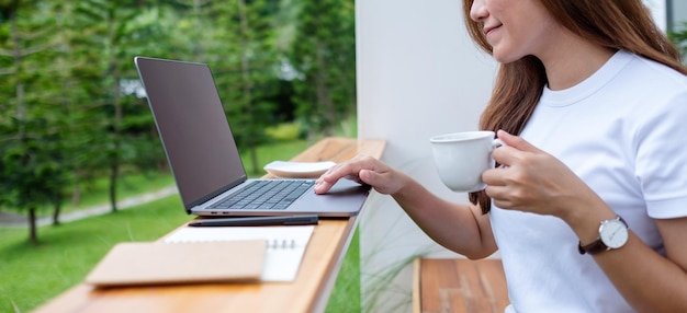 Closeup image of a beautiful young asian woman drinking coffee while using and working on laptop computer in the outdoors