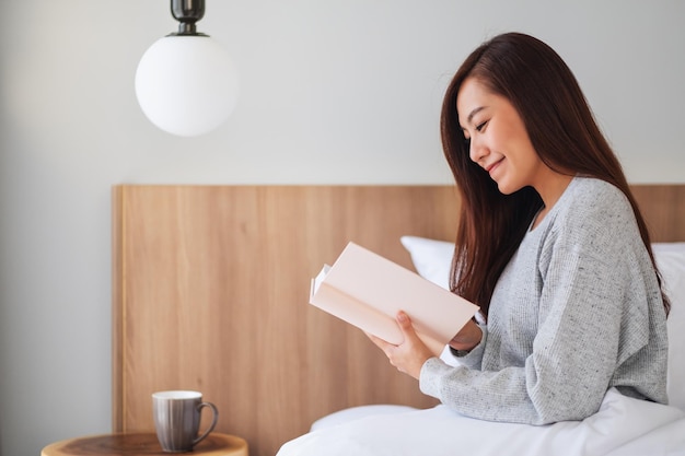 Closeup image of a beautiful young asian woman reading book in a white cozy bed at home