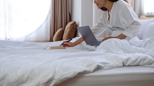 Closeup image of a beautiful young asian woman using laptop computer and working on paperwork while sitting on bed at home