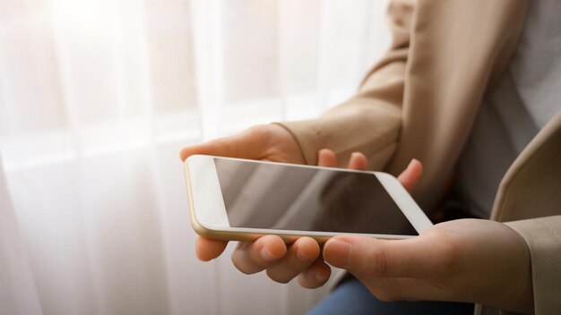 Closeup image Female in beige suit hands holding smartphone black screen mockup in bright room