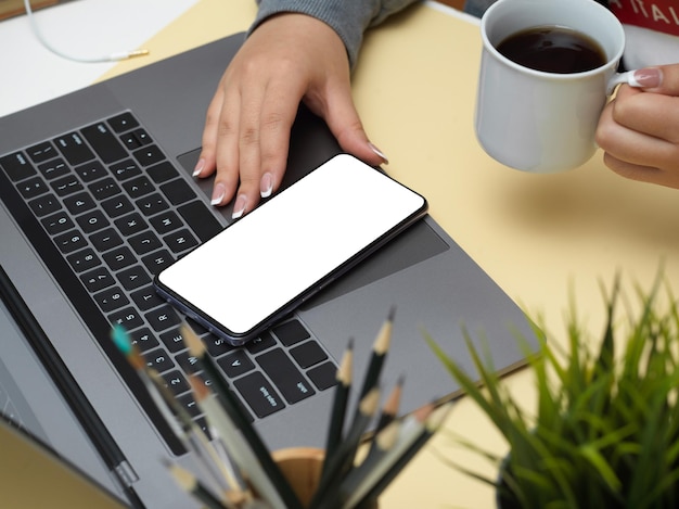 Closeup image Female holding black coffee mug and using smartphone over her laptop computer