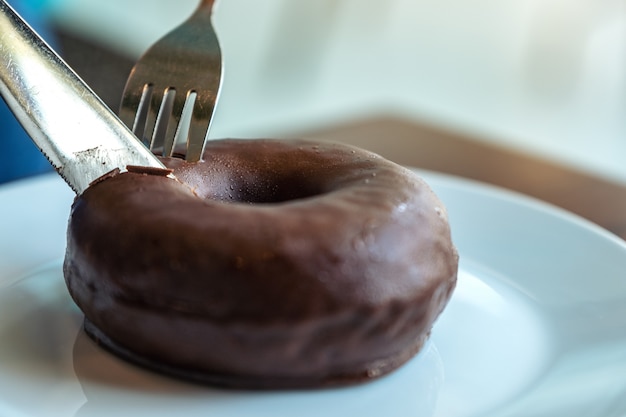 Closeup image of hands cutting a piece of chocolate donut by knife and fork for breakfast on wooden table