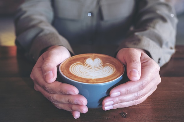 Closeup image of hands holding a blue cup of hot latte coffee with latte art on wooden table in cafe