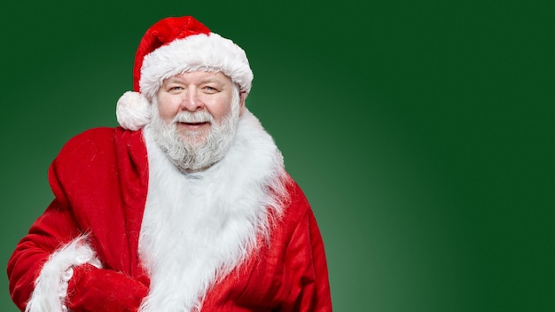 Closeup image of a happy laughing Santa Claus dressed in a red coat and a hat, keep a bag on his back, isolated green background. Copy space.