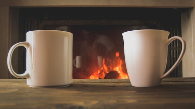 Closeup image of two cup of tea on wooden table next to burning fireplace