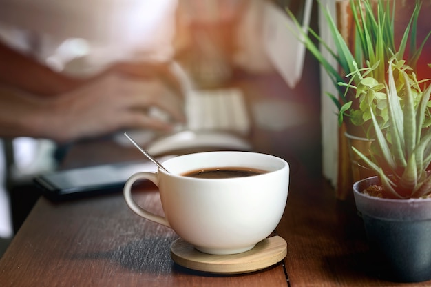 Closeup image of white cup of coffee on wooden teable with blur image of hand typing computer keyboard