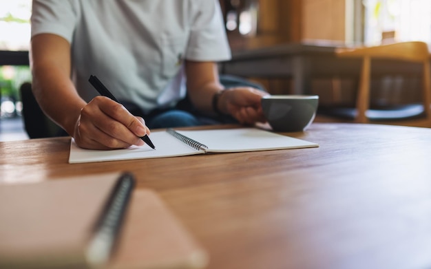 Closeup image of a woman drinking coffee and writing on a blank notebook on the table